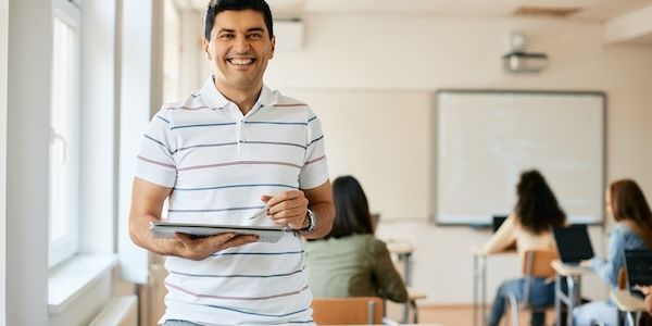 Happy high school teacher uses digital tablet during computer class and looking at camera.